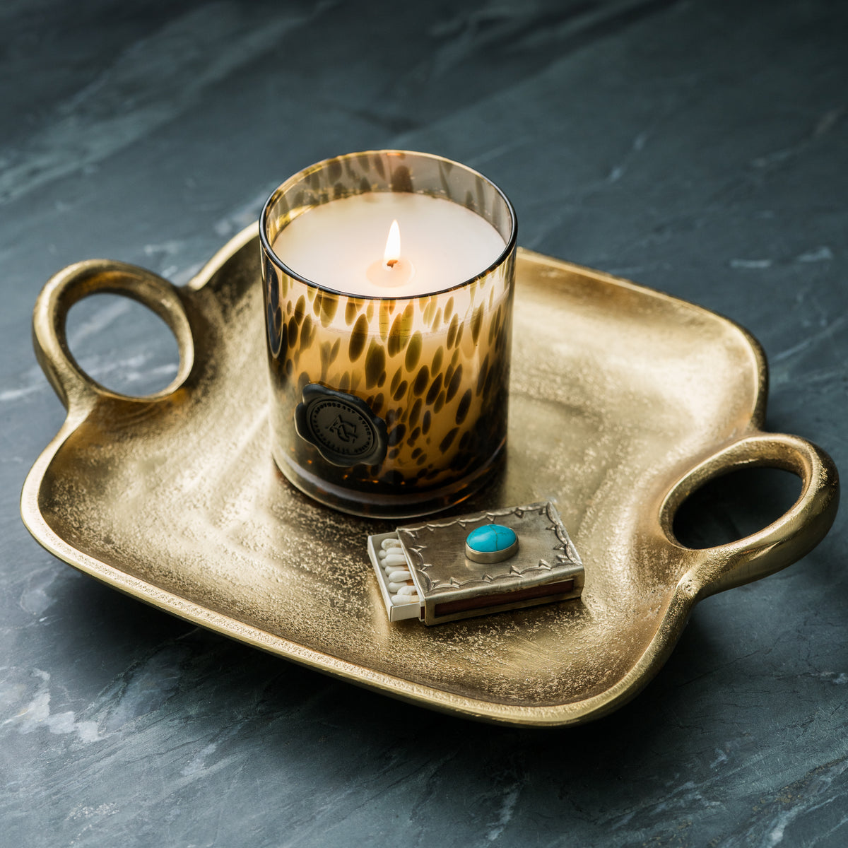Candle in a leopard print glass on a gold tray with a small decorative box on a dark surface