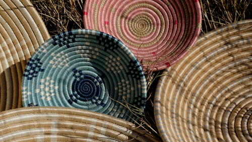 A group of hand-woven circular baskets in natural tan, pink, blue, and yellow-green tones, arranged on dry grass.